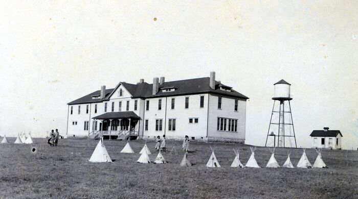 Historic Native American boarding school building with teepees and children on grassy field in early 1900s.