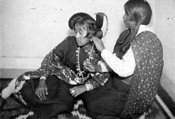 Two Native American girls in traditional clothing sitting on a patterned blanket at a boarding school in the 1910s.