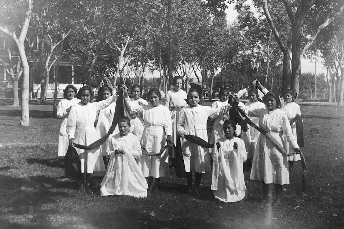 Native American girls in white dresses holding ribbons outdoors at a boarding school in the 1910s.