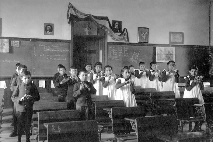 Native American children playing recorders in a 1910s boarding school classroom with wooden desks and chalkboards.