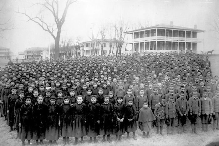 Large group of Native American children in uniform posing outside a boarding school building in the 1910s.
