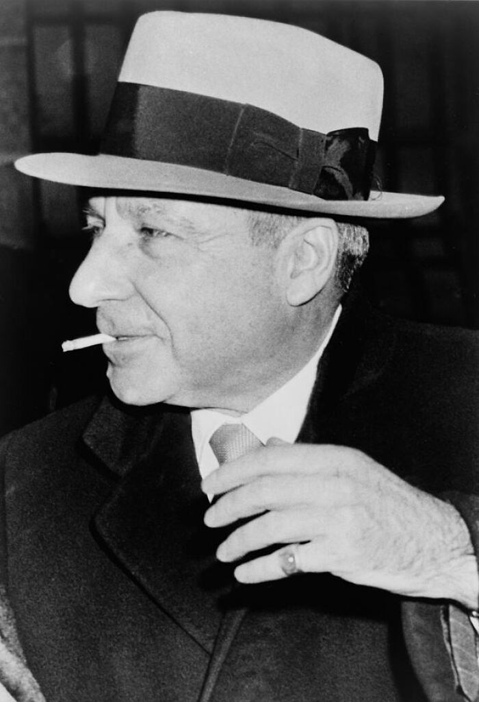 Black and white photo of a mob boss smoking a cigarette, wearing a fedora and suit, representing notorious mob bosses.