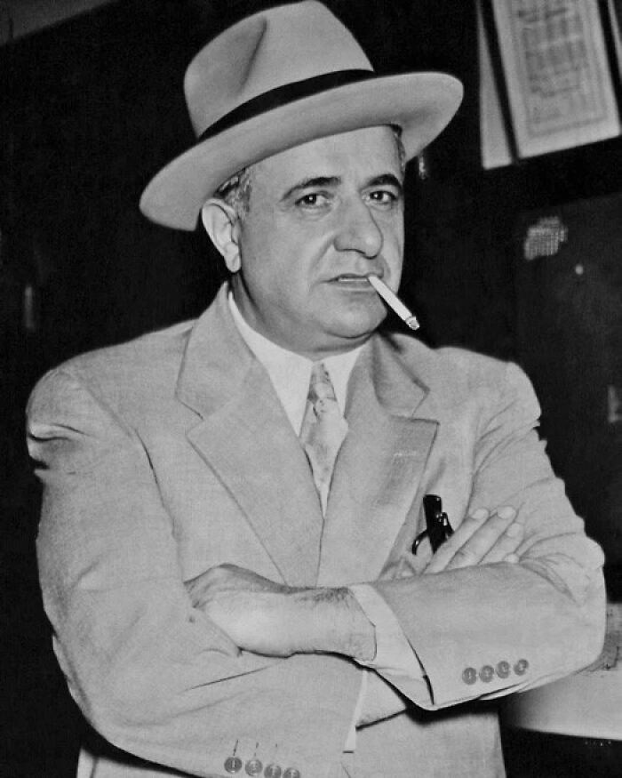 Black and white photo of a notorious mob boss wearing a hat and suit with a cigarette in his mouth, arms crossed.
