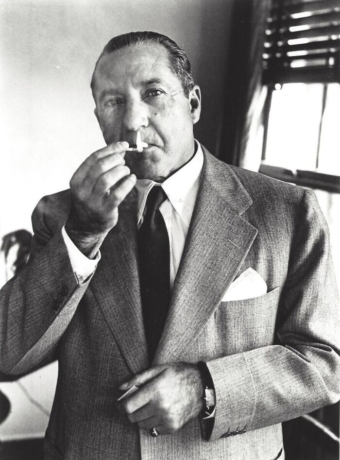 Black and white photo of a notorious mob boss in a suit lighting a cigarette by a window in a dimly lit room.