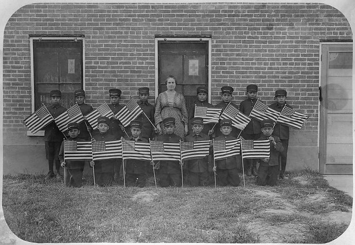 Group of Native American children holding American flags standing outside a boarding school in the 1910s.