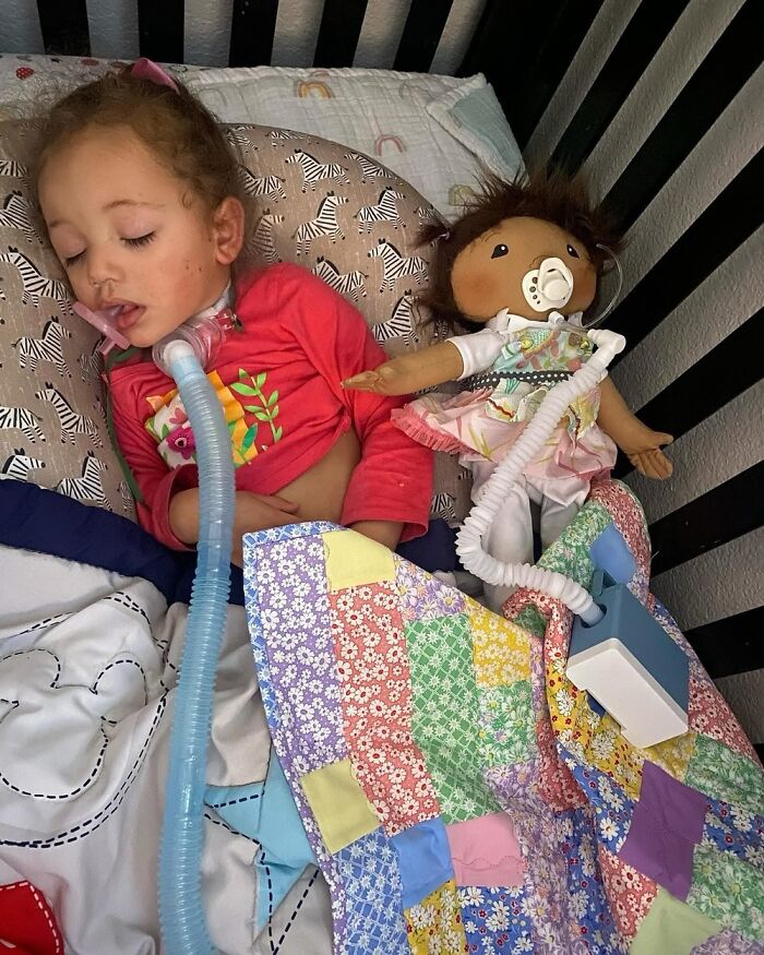 Child resting in bed with a medical ventilator and a doll designed to represent her rare health condition.