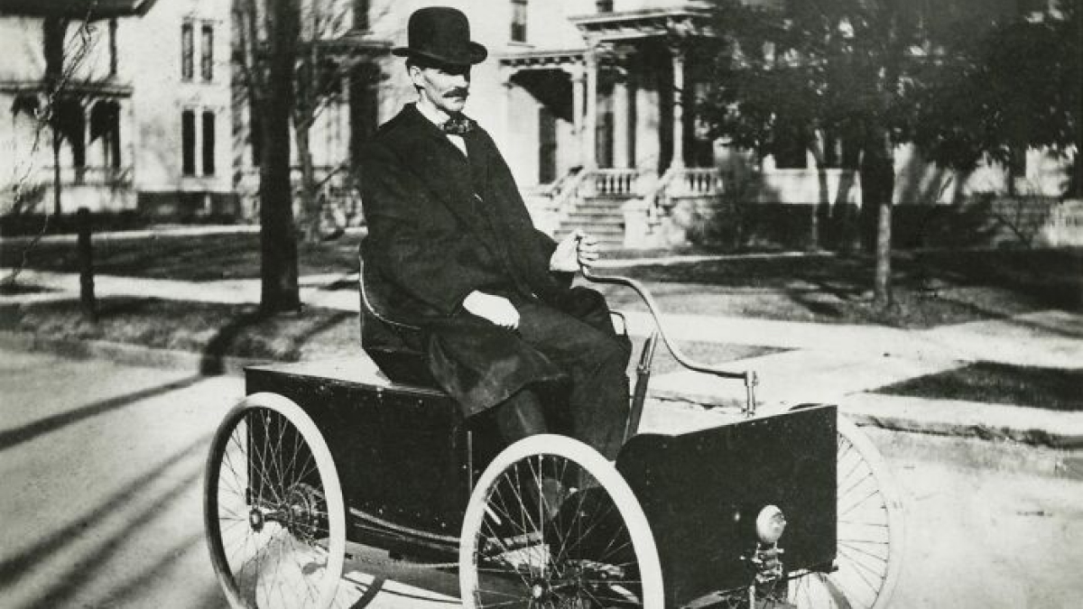 Man in 1800s America wearing a bowler hat and coat riding an early motorized car on a residential street.