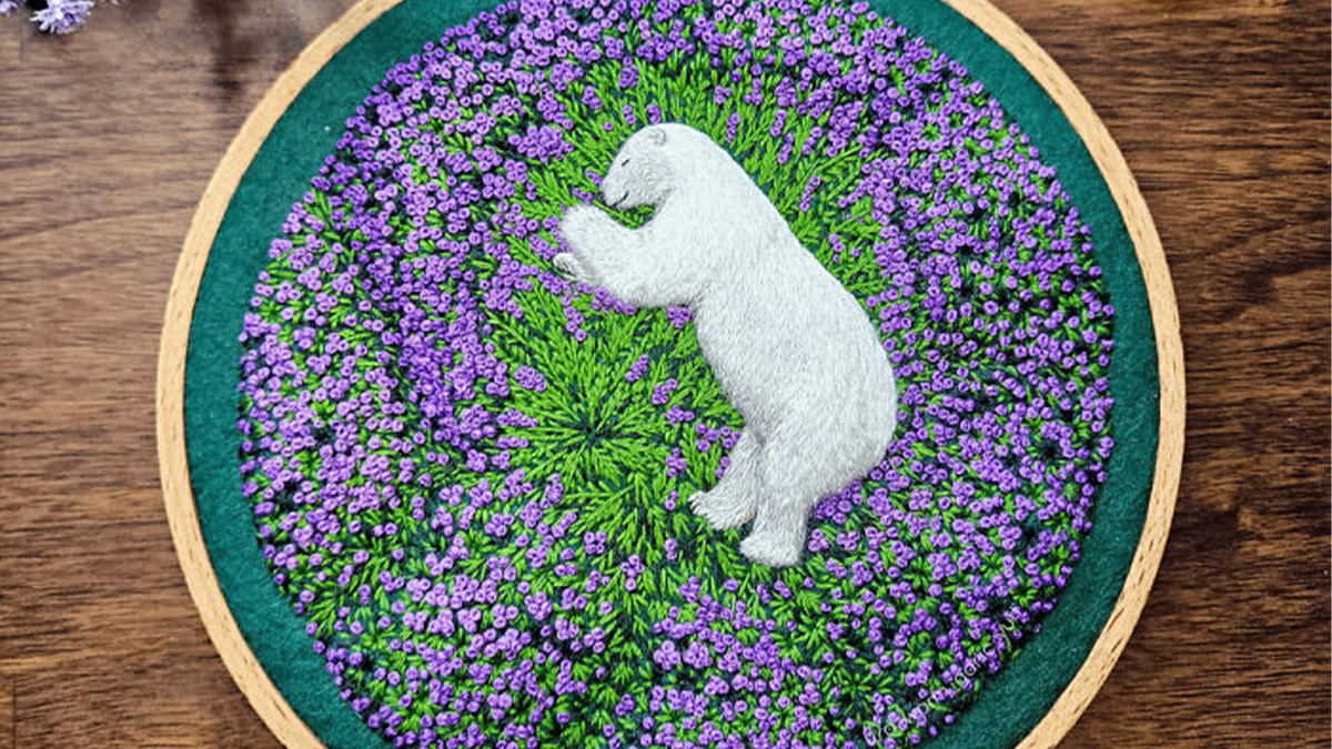 Embroidery hoop art showing a white bear surrounded by dense purple flowers and green leaves, showcasing embroidery skills.