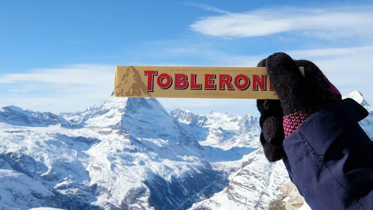 Hand in winter gloves holding Toblerone chocolate with snowy Swiss mountains in the background showcasing world geography.