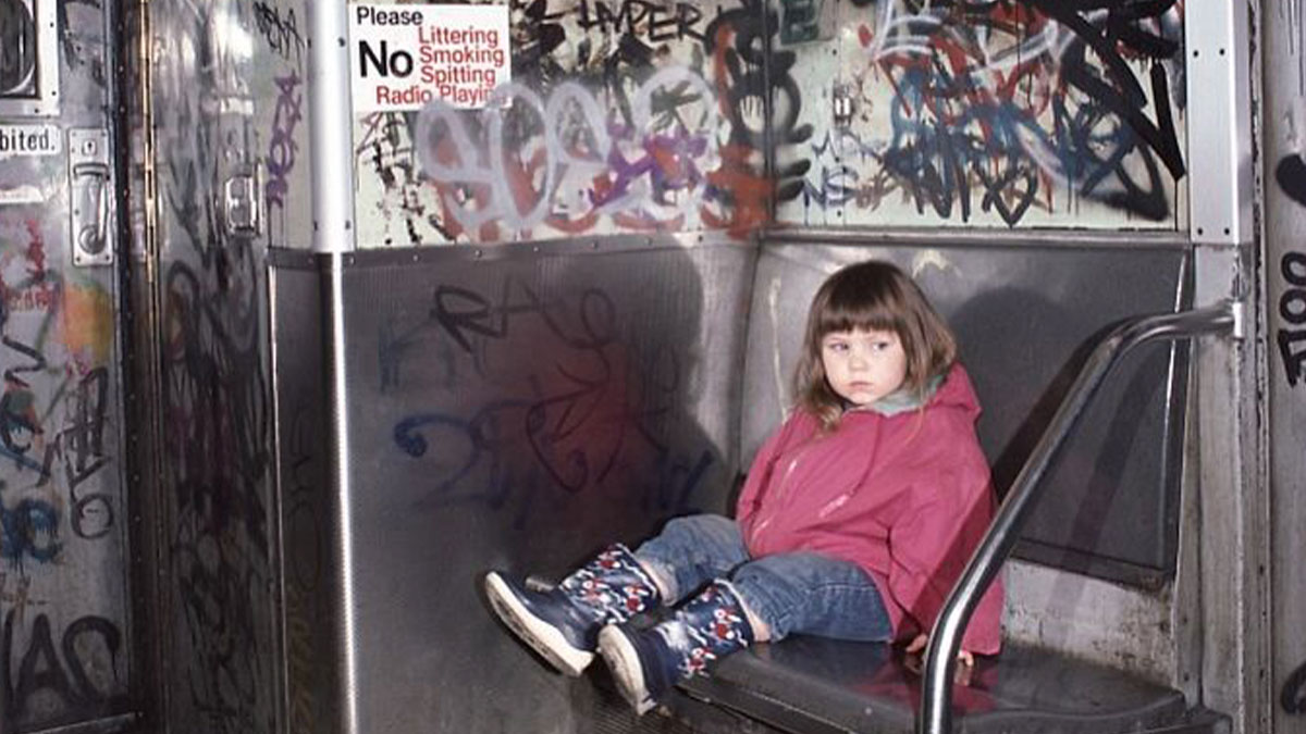 Young girl in a pink jacket sitting on a metal subway seat surrounded by graffiti in a historical photo.