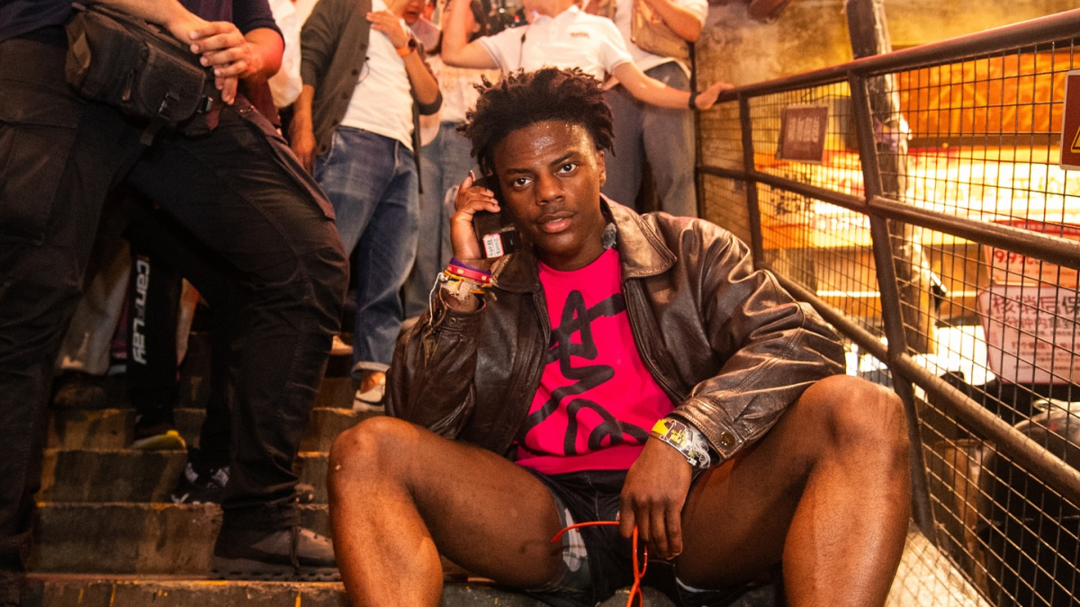 Ishowspeed net worth growth shown by young man sitting on stairs in leather jacket and red shirt using a phone amid crowd
