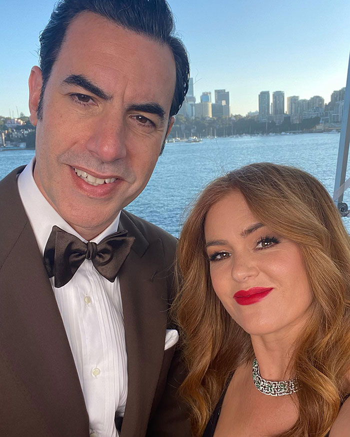 Isla Fisher and Sasha Baron Cohen smiling together outdoors with city skyline and water in the background. Isla Fisher and Sasha Baron Cohen smiling together outdoors with city skyline and water in the background.