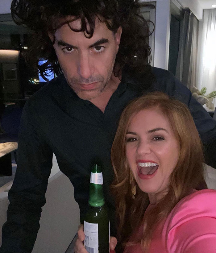 Isla Fisher smiling and holding a bottle, with Sasha Baron Cohen standing behind her in a casual indoor setting. Isla Fisher smiling and holding a bottle, with Sasha Baron Cohen standing behind her in a casual indoor setting.