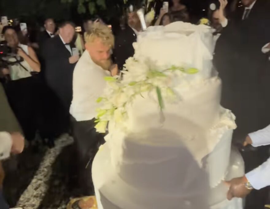 Jake Paul smashing a large wedding cake at a crowded event with people taking photos in the background Jake Paul smashing a large wedding cake at a crowded event with people taking photos in the background