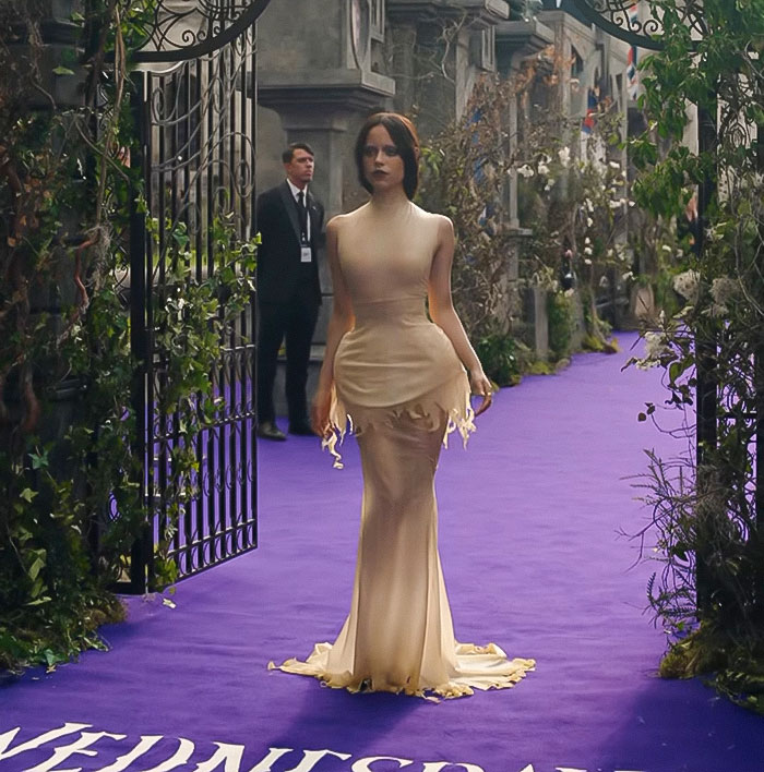 Jenna Ortega wearing a sleek beige gown on a purple carpet at an event, showcasing Wednesday looks in fashion. Jenna Ortega wearing a sleek beige gown on a purple carpet at an event, showcasing Wednesday looks in fashion.