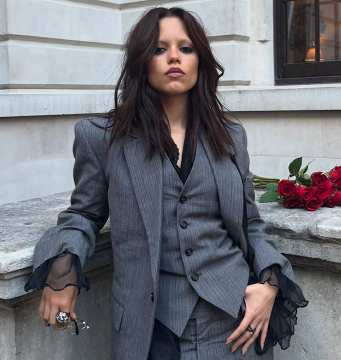 Jenna Ortega posing in a gray pinstripe suit with black lace cuffs, leaning against a stone ledge with red roses nearby. Jenna Ortega posing in a gray pinstripe suit with black lace cuffs, leaning against a stone ledge with red roses nearby.