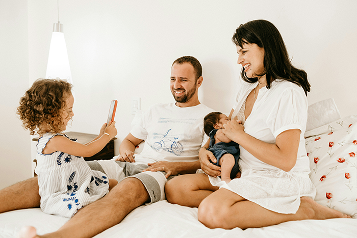Family moment with a smiling woman holding a baby while a man and child enjoy taking photos in a cozy bedroom setting.