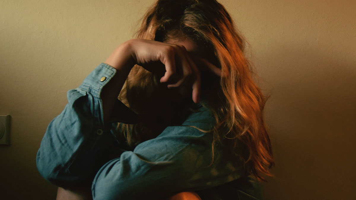 Woman covering face sitting in dim light, conveying frustration and emotion related to sexism and job challenges.