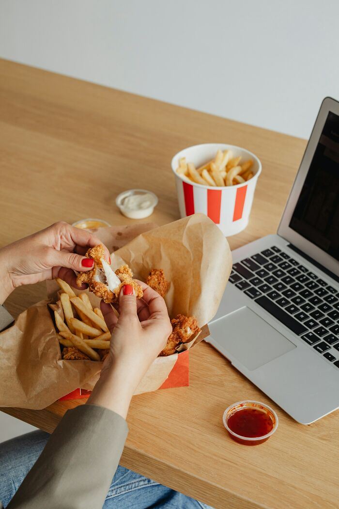 Hands holding fried chicken with fries and sauces next to a laptop, illustrating PR campaigns and decisions fails concept.