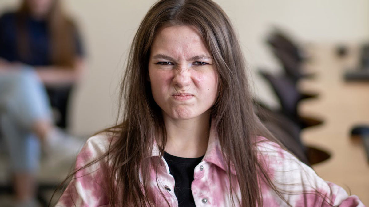 Teen girl with long brown hair and a pink shirt showing an angry facial expression in a classroom setting.