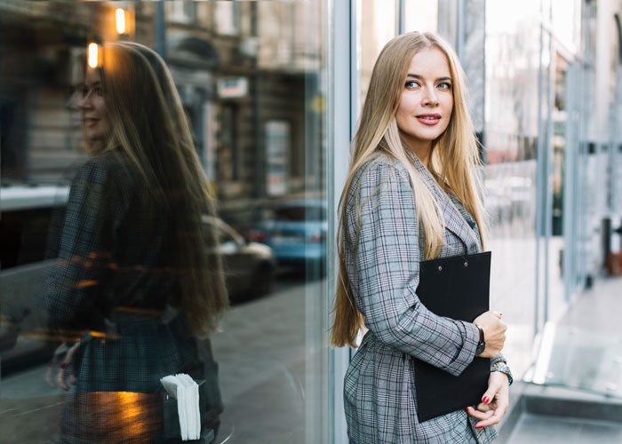 Blonde woman in plaid blazer standing by glass window, holding clipboard, looking away on urban street scene.