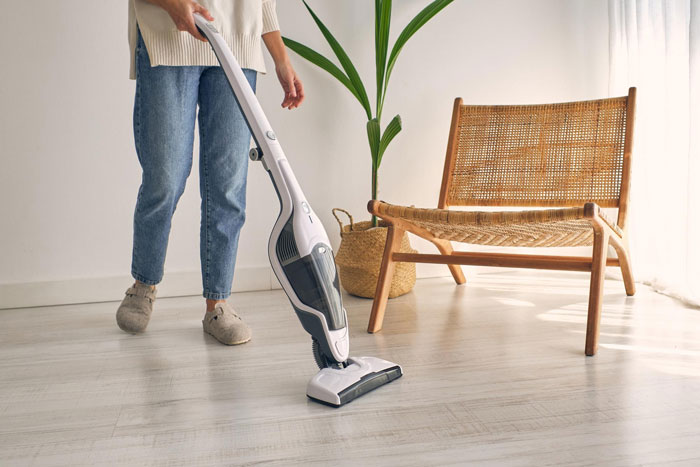 Person using a modern vacuum cleaner on light wood floor near wooden chair and potted plant in bright room. Person using a modern vacuum cleaner on light wood floor near wooden chair and potted plant in bright room.