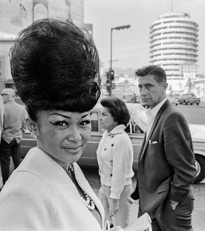 LA woman with beehive hairstyle, pearl necklace, and white coat walking city streets with vintage cars in background.