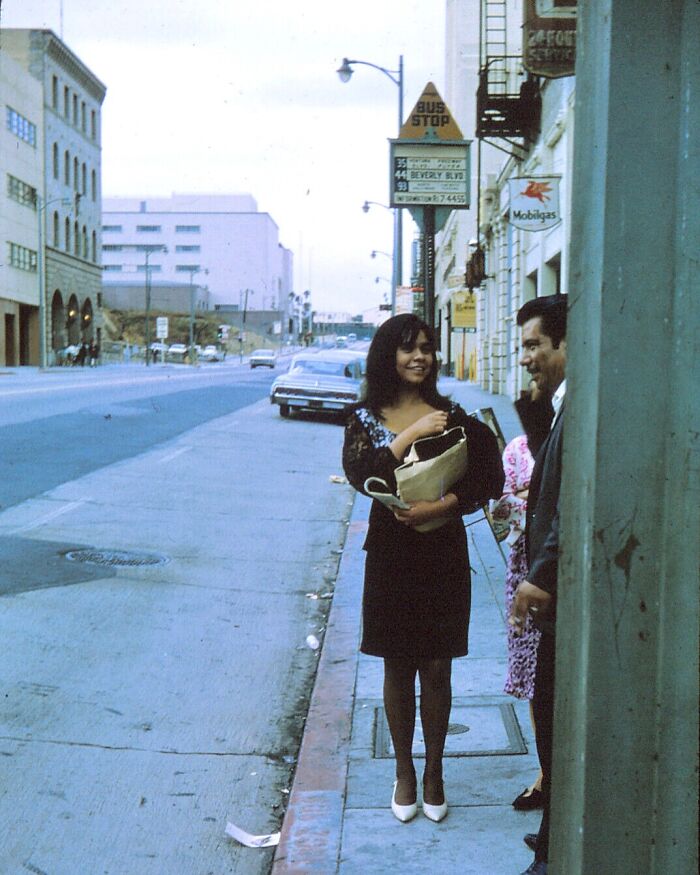 LA woman smiling and holding bags while standing on a sidewalk near a bus stop in a 1960s urban street scene.