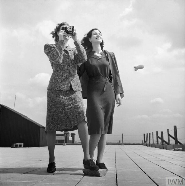 Two British women on a rooftop during WWII, showcasing style while one photographs and a blimp floats in the sky.
