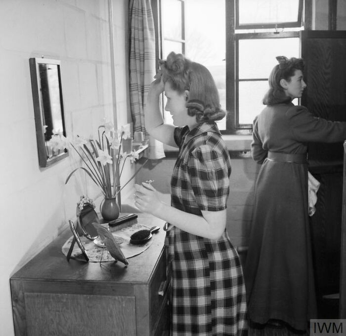 Two British women in 1940s dresses applying makeup and styling hair, showcasing British women style during WWII restrictions.