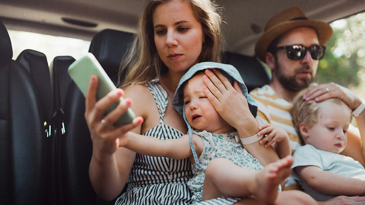 Wife with two young children looking at phone while husband with man-child behavior sits beside them on family vacation.