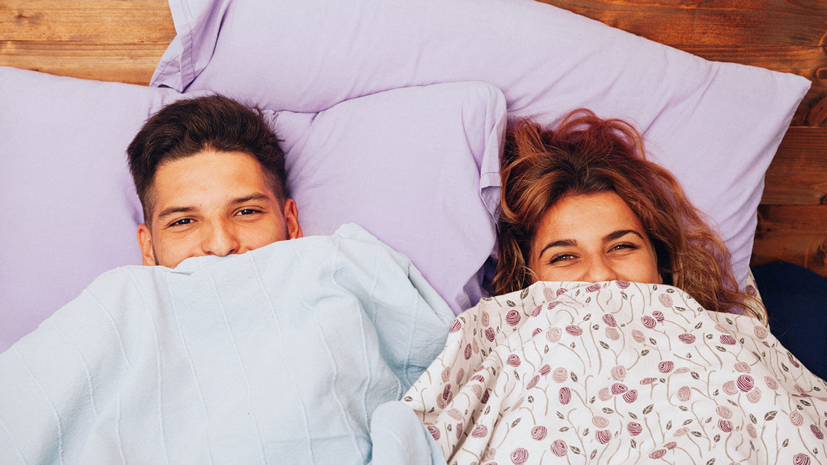 Couple adopting cool habits after visiting other countries, smiling while lying in bed under colorful blankets.