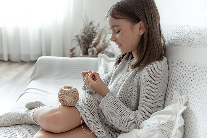 Young girl knitting with yarn while sitting on a white sofa in a cozy living room setting. Young girl knitting with yarn while sitting on a white sofa in a cozy living room setting.