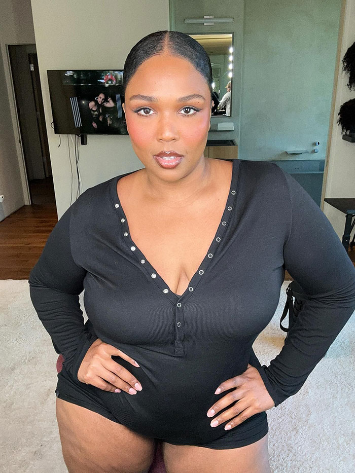 Lizzo posing confidently indoors wearing a black long sleeve outfit with a serious expression and slicked-back hair. Lizzo posing confidently indoors wearing a black long sleeve outfit with a serious expression and slicked-back hair.