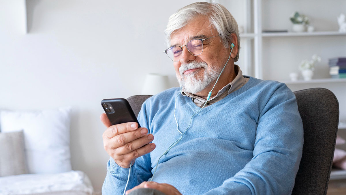 Elderly man in blue sweater relaxing and enjoying content on smartphone with earphones, illustrating low effort high reward.