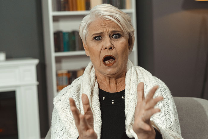 Older woman with short white hair looks shocked and upset while gesturing with her hands in a home setting Older woman with short white hair looks shocked and upset while gesturing with her hands in a home setting