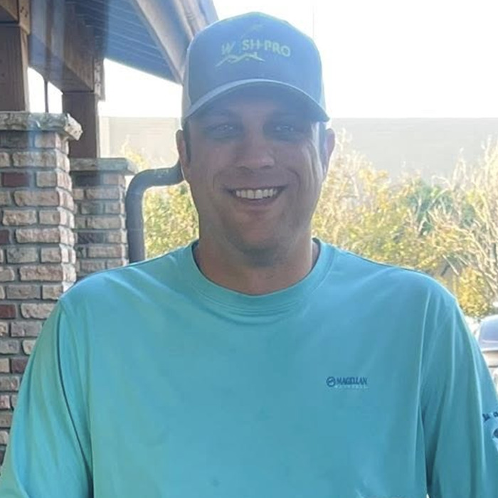 Man wearing a blue shirt and cap, smiling outdoors with sunlight and brick wall in the background, related to breast milk assault case. Man wearing a blue shirt and cap, smiling outdoors with sunlight and brick wall in the background, related to breast milk assault case.