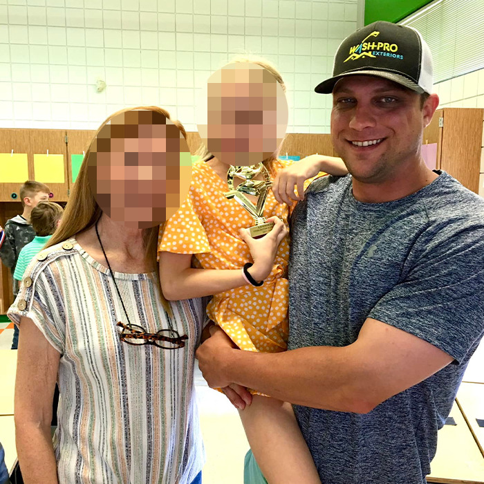 Man wearing a black cap holding a child in a yellow dress with an adult woman nearby in a casual indoor setting. Man wearing a black cap holding a child in a yellow dress with an adult woman nearby in a casual indoor setting.