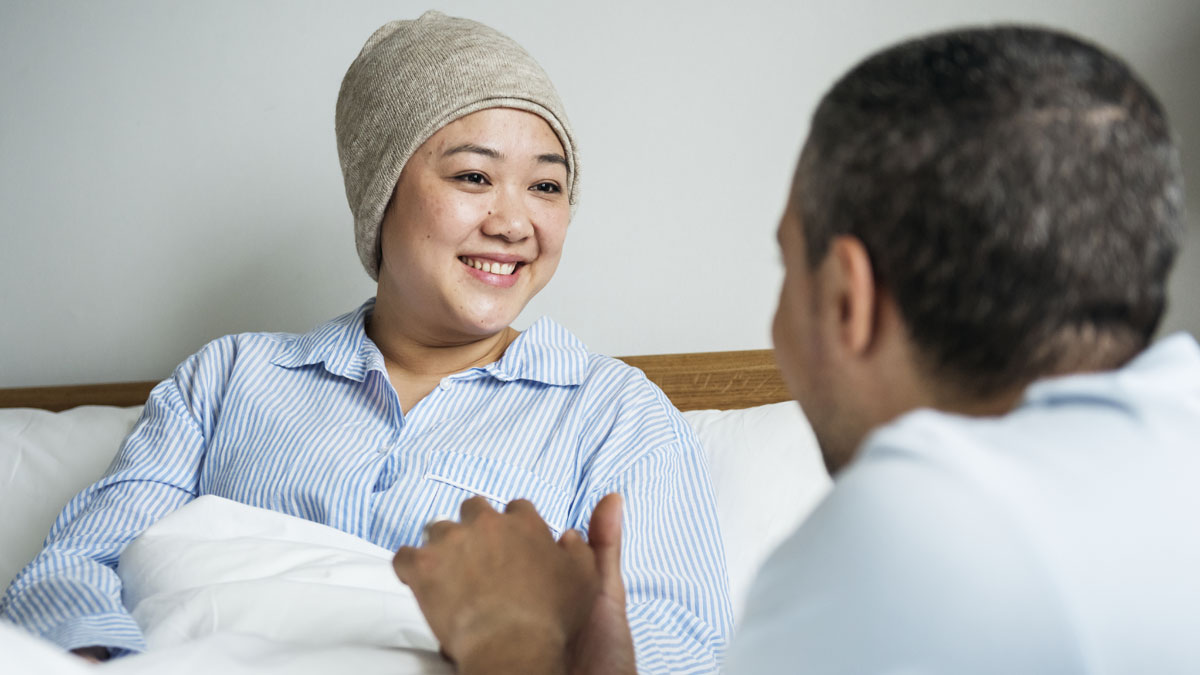 Woman in hospital bed wearing head scarf smiling at man holding her hands, reflecting marriage and terminally ill care issues