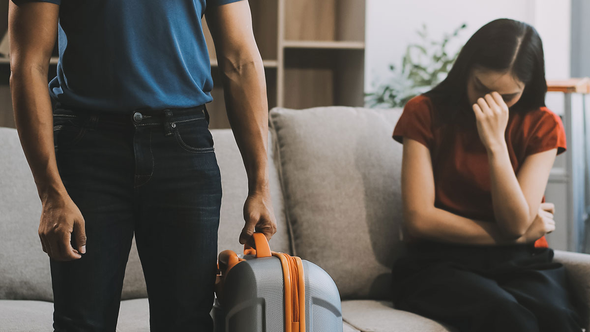 Woman upset on couch as boyfriend stands holding suitcase, highlighting trust issues in couples trip situation.