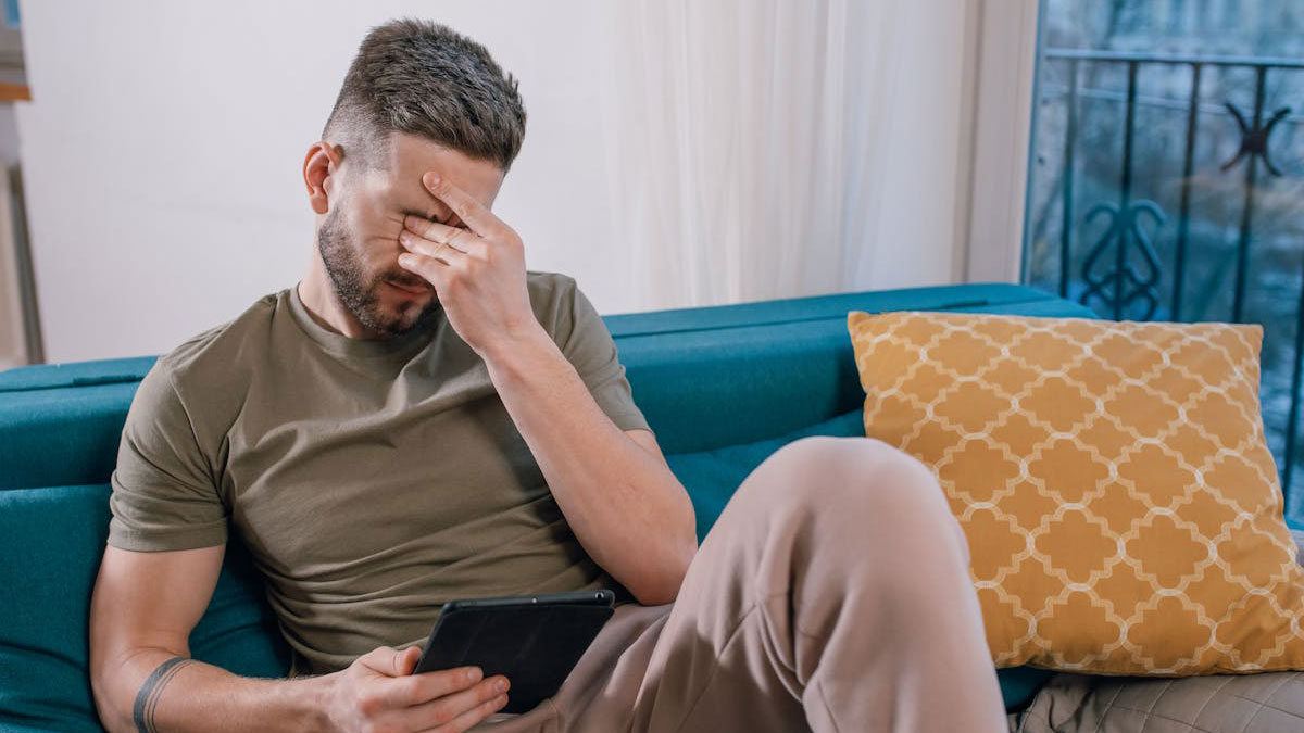 Man defends his wife, looks stressed while sitting on couch holding tablet, conflicted about cutting off parents.