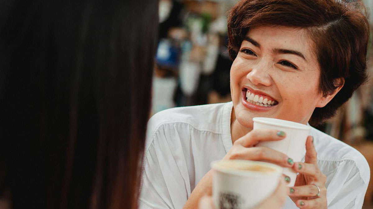 Woman smiling and holding a cup while chatting with a friend, illustrating tiredness of being used as an ATM.
