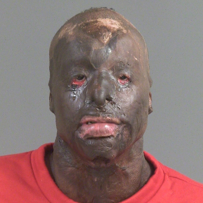 Man resembling Batman villain with extensive facial tattoos and scars wearing a red shirt posing for mugshot photo. Man resembling Batman villain with extensive facial tattoos and scars wearing a red shirt posing for mugshot photo.