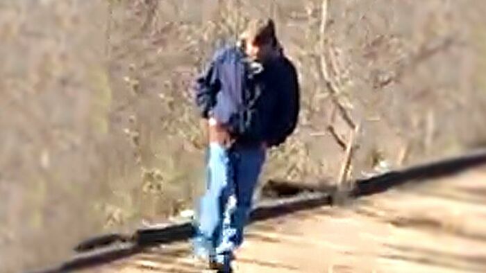 A man in a dark jacket and blue jeans walking on a path with bare trees in the background, context unclear.