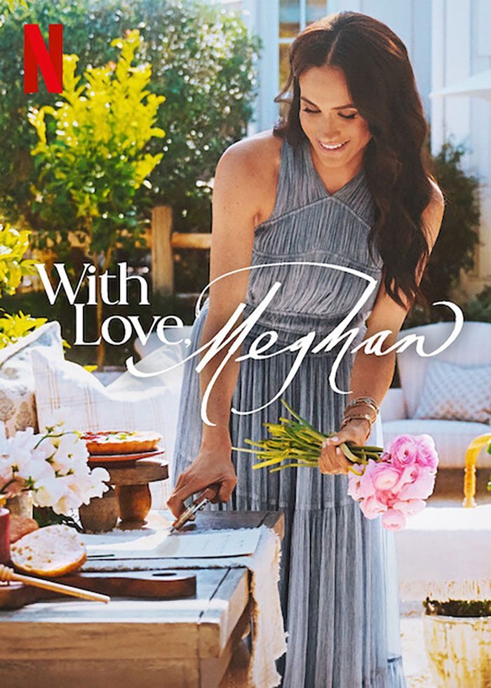 Meghan Markle in a grey dress holding flowers near a table of food in an outdoor setting promoting her cooking show. Meghan Markle in a grey dress holding flowers near a table of food in an outdoor setting promoting her cooking show.