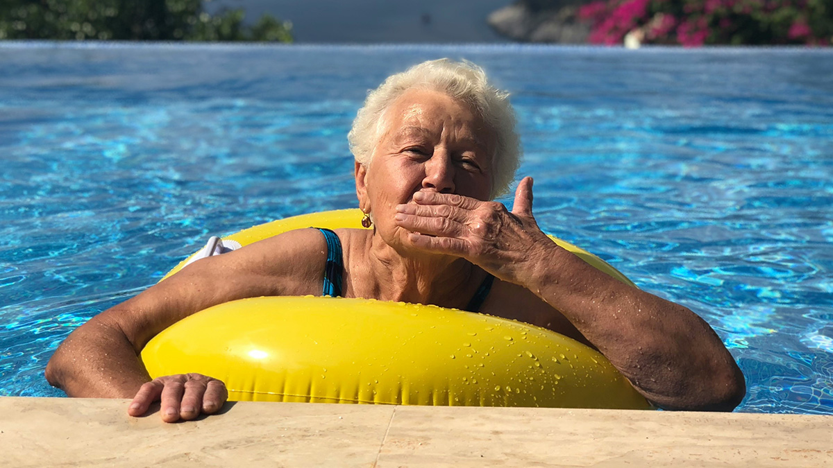 Elderly woman in swimming pool with yellow float, sending a kiss, symbolizing stalker MIL at a coupleu2019s vacation.