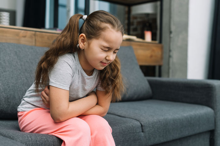 Young girl sitting on a couch holding her stomach in pain, depicting lactose intolerance symptoms in children. Young girl sitting on a couch holding her stomach in pain, depicting lactose intolerance symptoms in children.