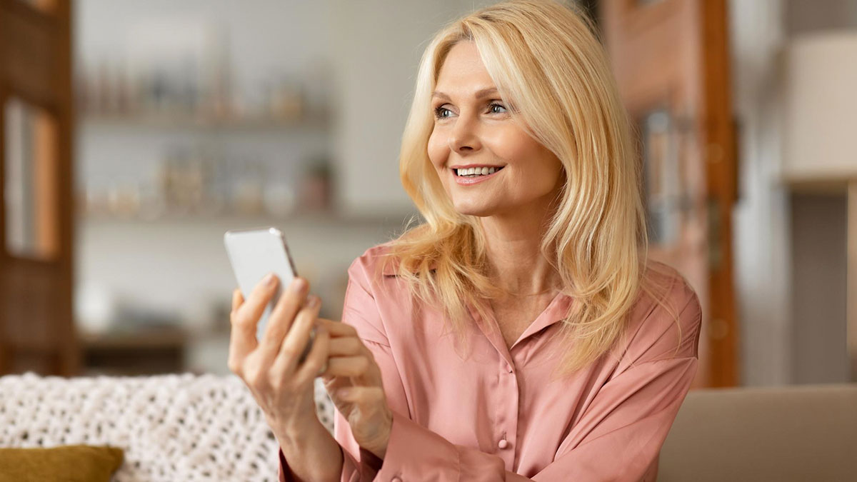 Middle-aged woman smiling and holding smartphone, reflecting on not moving on after MIL live streamed her sleeping.