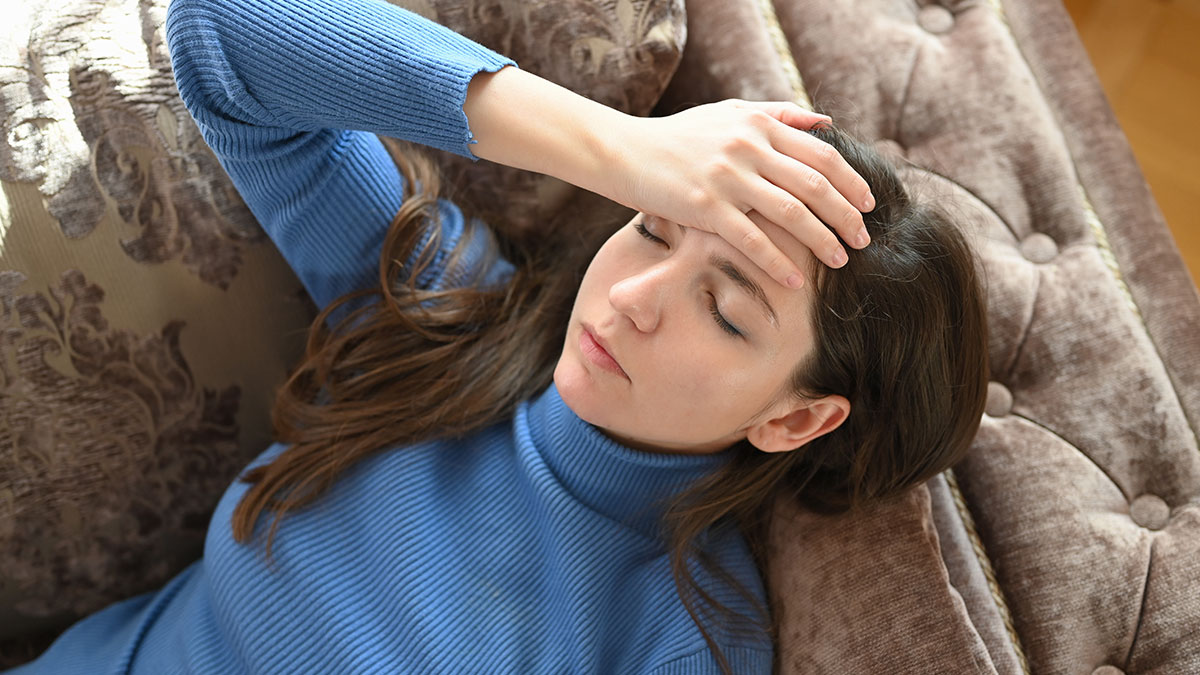 Woman in blue sweater lying on couch with hand on forehead, appearing unwell and stressed from poisoning accusations.