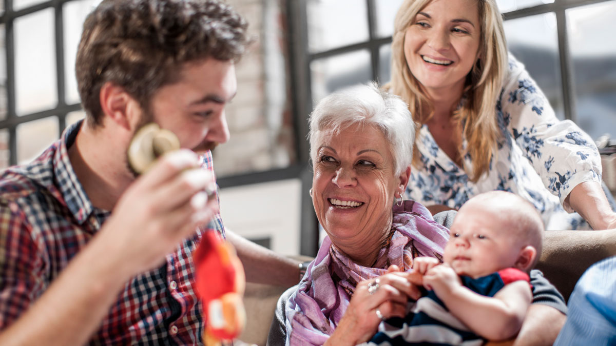 Woman smiling while holding baby, interacting with man and younger woman in a bright living room setting, family moment.